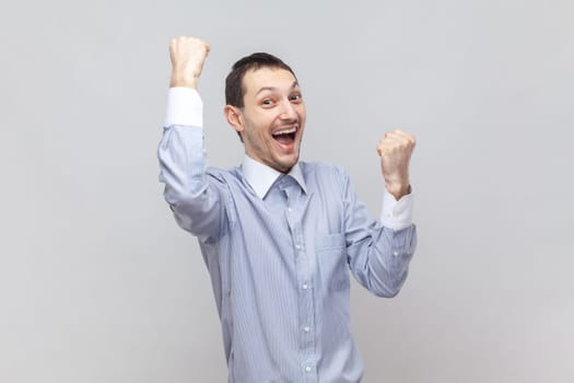 Portrait of overjoyed happy successful man standing with raised clenched fists, screaming with happiness, celebrating victory, wearing light blue shirt. Indoor studio shot isolated on gray background.