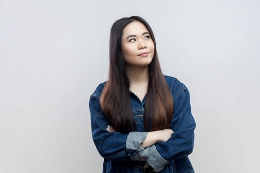 Portrait of pensive thoughtful young adult brunette woman in blue denim jacket standing with crossed arms, looking away, thinking, Indoor studio shot isolated on gray background.