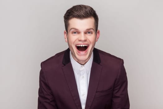 Portrait of handsome man laughs at something funny, looking at camera, having fun, expressing positive emotions, wearing violet suit and white shirt. Indoor studio shot isolated on grey background.