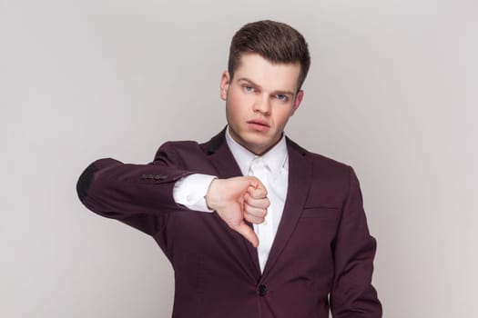 Portrait of handsome man shows disapproval sign, keeps thumb down, expresses dislike, frowns face in discontent, wearing violet suit and white shirt. Indoor studio shot isolated on grey background.