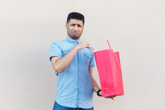 Portrait of sad stressed depressed man wearing denim shirt being disappointed of his shopping in mall, holding paper bag and crying. Indoor studio shot isolated on gray background.