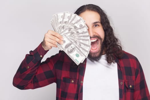 Portrait of amazed surprised excited bearded man with long curly hair in checkered red shirt covering half of face with dollar banknotes. Indoor studio shot isolated on gray background.