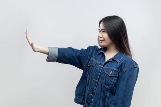 Portrait of brunette woman in denim jacket standing making stop gesture showing palm of hand, conflict prohibition warning about danger, stop bullying. Indoor studio shot isolated on gray background.