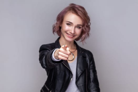 Portrait of smiling satisfied woman with short hairstyle standing pointing fore finger to camera, choosing and needs you, wearing black leather jacket. Indoor studio shot isolated on grey background.