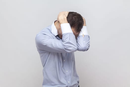 Portrait of despair sad upset man standing and hiding his face with hand, having problems, avoids very loud noise, wearing light blue shirt. Indoor studio shot isolated on gray background.