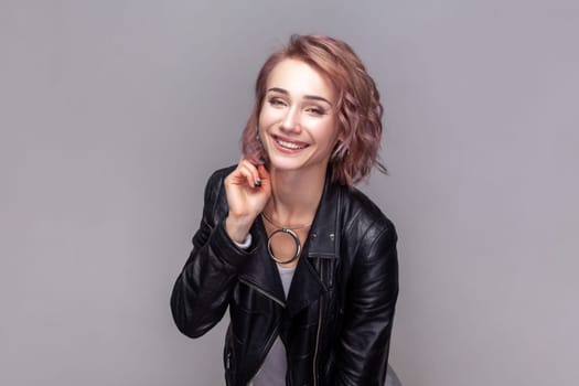 Portrait of charming beautiful adorable woman with short hairstyle standing looking at camera with toothy smile, wearing black leather jacket. Indoor studio shot isolated on grey background.