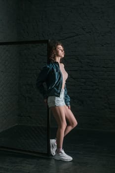 Full length profile portrait of beautiful young adult woman with wavy hair standing near fence on dark brick wall background, looking away with serious expression. Indoor studio shot.