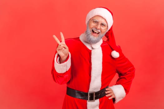 Amazed elderly man with gray beard wearing santa claus costume standing with raised hands and showing v sign or peace, victory gesture. Indoor studio shot isolated on red background.