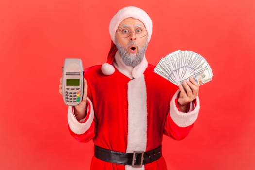 Portrait of shocked elderly man with gray beard wearing santa claus costume standing with open mouth, holding terminal and fan of dollars banknotes. Indoor studio shot isolated on red background.