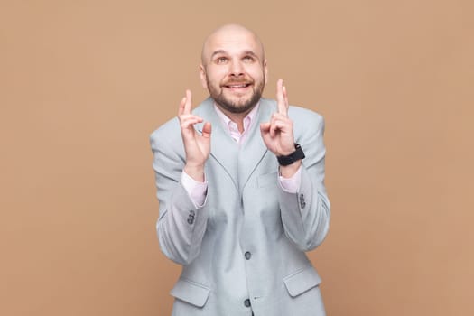 Portrait of hopeful nervous bald bearded man crossing fingers for luck, hope for better, ritual, looks up with hopeful look, wearing gray jacket. Indoor studio shot isolated on brown background.