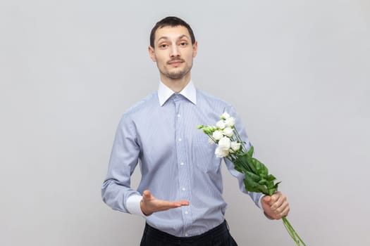 Portrait of handsome young adult man standing with bouquet of flowers, showing beautiful plants, gift for anniversary, wearing light blue shirt. Indoor studio shot isolated on gray background.
