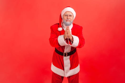 Forgive me or give me one chance more. Elderly man with gray beard wearing santa claus costume standing with palm hands, looking and begging. Indoor studio shot isolated on red background.