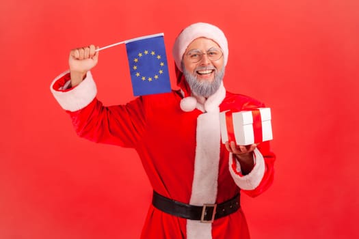 Portrait of smiling elderly man with gray beard wearing santa claus costume holding wrapped present box and waving Europe union flag, looks at camera. Indoor studio shot isolated on red background.