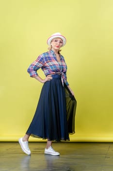 Full length portrait of mature woman wearing hat and eyeglasses standing, posing and looking at camera, having serious expression, keeps hand on hip. Indoor studio shot isolated on yellow background