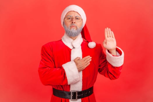 Serious elderly man with gray beard wearing santa claus costume standing raising hand and saying swear, making loyalty oath, pledging allegiance. Indoor studio shot isolated on red background.