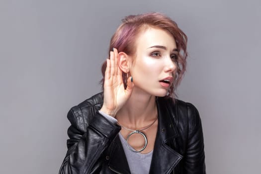 Portrait of attentive adorable woman with short hairstyle standing with hand near ear, listening private conversation, wearing black leather jacket. Indoor studio shot isolated on grey background.