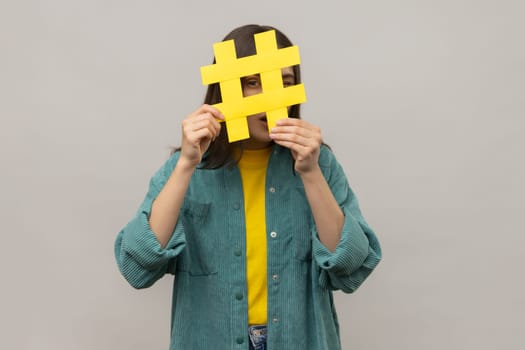 Portrait of curious woman looking through yellow hashtag sign, looking for proper posts in social media, spying, wearing casual style jacket. Indoor studio shot isolated on gray background.