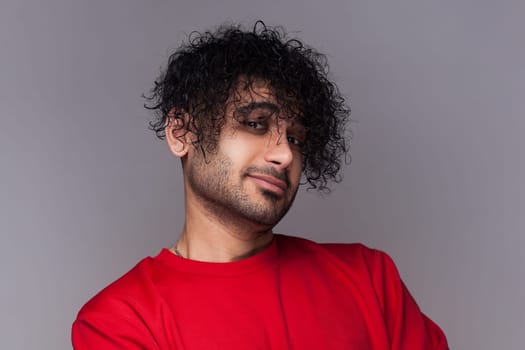 Portrait of confident proud satisfied handsome bearded man with curly hair, looking at camera with smile, wearing red jumper. Indoor studio shot isolated on gray background.