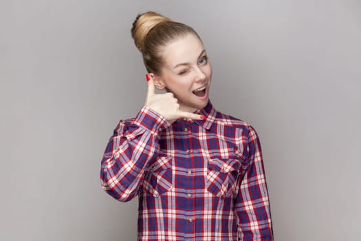 Portrait of beautiful positive woman with bun hairstyle flirting with attractive guy, showing call me gesture, wearing checkered shirt. Indoor studio shot isolated on gray background.