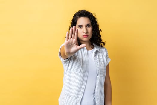 Portrait of woman with dark wavy hair showing stop gesture with palm of hand, trying to stop abuser, rejection, afraid of domestic violence. Indoor studio shot isolated on yellow background.