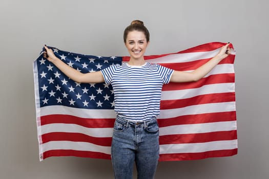 Portrait of joyful young adult smiling woman wearing striped T-shirt holding big american flag, expressing positive emotions and happiness. Indoor studio shot isolated on gray background.