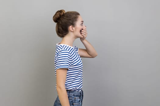 Side view of astonished woman wearing striped T-shirt covering mouth with hand, afraid to say secret, child terrified to speak. Indoor studio shot isolated on gray background.