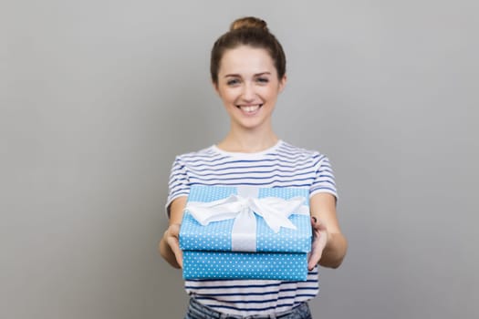 Portrait of woman wearing striped T-shirt holding out present box, giving gift, looking at camera with optimistic expression, greeting. Indoor studio shot isolated on gray background.