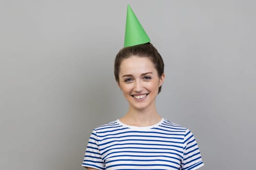 Portrait of smiling positive delighted woman wearing striped T-shirt and party cone, looking at camera with toothy smile. Indoor studio shot isolated on gray background.