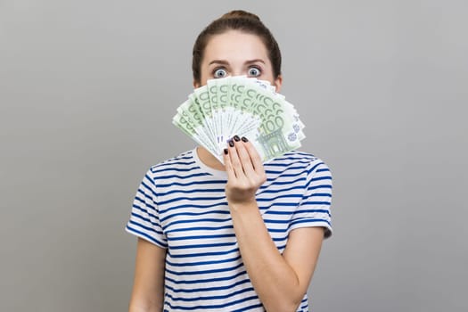 Portrait of surprised woman wearing striped T-shirt peeping out euro banknotes with exited look, big jackpot, financial success. Indoor studio shot isolated on gray background.