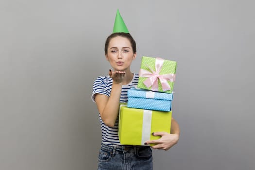 Portrait of flirting woman wearing striped T-shirt and party cone, holding stack of present boxes, sending air kiss. Indoor studio shot isolated on gray background.