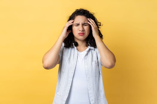 Portrait of woman with dark wavy hair frowning and clasping sore head, suffering intense headache, having migraine, fever symptoms. Indoor studio shot isolated on yellow background.