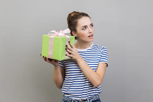 Portrait of woman wearing striped T-shirt shaking wrapped present box, being interested what inside, looking away with curious facial expression. Indoor studio shot isolated on gray background.