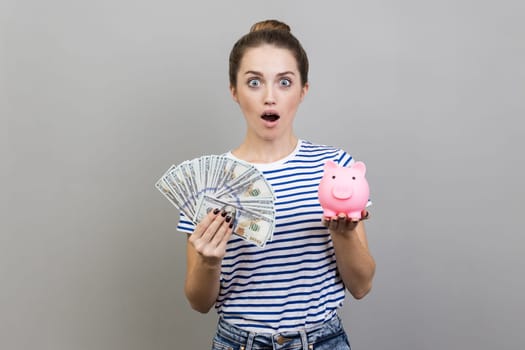 Portrait of astonished surprised woman wearing striped T-shirt holding fan of dollar bills and piggy bank, savings, growth of deposit. Indoor studio shot isolated on gray background.
