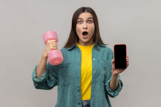 Portrait of amazed surprised holding pink dumbbell and blank screen smartphone in hands, satisfied with sports app, wearing casual style jacket. Indoor studio shot isolated on gray background.