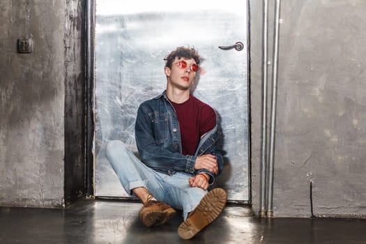 Full length portrait of calm attractive relaxed fashionable model man in bright red sunglasses and denim casual style, sitting near metallic door, looking at camera. Indoor studio shot.