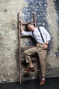 Portrait of trendy stylish young model man wearing white shirt and beige pants posing and lean on old wooden ladder on brick wall background. Indoor studio shot.