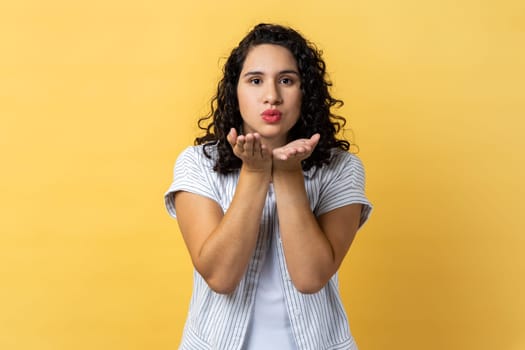 Woman with dark wavy hair sending air kiss to camera, flirting and demonstrating love affection feelings, keeps eyes closed, romantic relationships. Indoor studio shot isolated on yellow background.