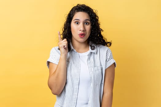 Portrait of excited amazed woman with dark wavy hair pointing upwards, looking at camera with open mouth, having new great idea. Indoor studio shot isolated on yellow background.