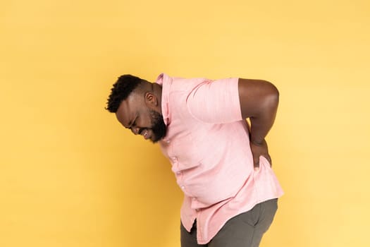 Portrait of exhausted unhealthy man wearing pink shirt touching aching back, suffering lower lumbar discomfort, muscle pain, injured spine disk. Indoor studio shot isolated on yellow background.