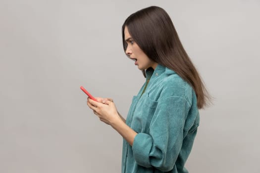 Side view of shocked pretty woman reading message on smartphone with open mouth, using mobile device for communication, wearing casual style jacket. Indoor studio shot isolated on gray background.