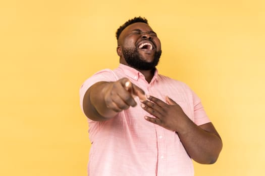 Portrait of funny positive bearded young adult man wearing pink shirt laughing out loud holding belly and pointing finger on you, mockery. Indoor studio shot isolated on yellow background.