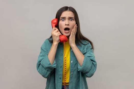 Amazed woman holding phone handset surprised by conversation, open mouth shocked by unbelievable news, discussing rumors on telephone. Indoor studio shot isolated on gray background.