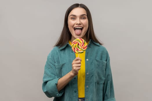 Childish attractive woman licking multicolor candy, wants to eat, looking at camera, showing tongue out, wearing casual style jacket. Indoor studio shot isolated on gray background.