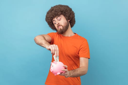 Portrait of man with Afro hairstyle wearing orange T-shirt investing, putting dollar banknote into piggy bank, proud with money savings. Indoor studio shot isolated on blue background.