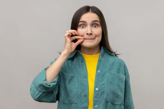 Portrait of attractive woman with mystery look making zip gesture to close mouth, keeping secret, zipping lips, wearing casual style jacket. Indoor studio shot isolated on gray background.