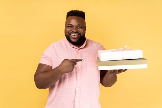 Portrait of delighted positive cheerful man wearing pink shirt holding and pointing at gift boxes, looking at camera with happy smile. Indoor studio shot isolated on yellow background.