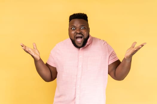 What a surprise. Portrait of man wearing pink shirt standing with wide open mouth and raising hands in amazement, shocked by unbelievable success. Indoor studio shot isolated on yellow background.