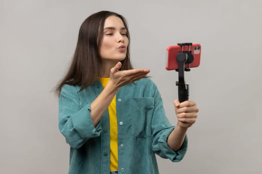 Romantic woman holding smartphone on steadycam in hand, blogger broadcasting livestream or making selfie, sending air kiss, wearing casual style jacket. Indoor studio shot isolated on gray background