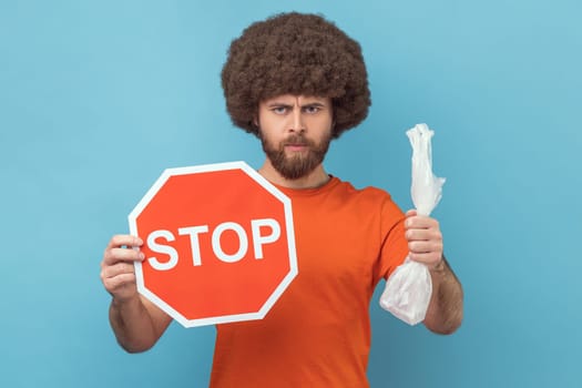 Portrait of serious responsible man with Afro hairstyle wearing orange T-shirt holding red stop sign and plastic package, looking at camera. Indoor studio shot isolated on blue background.