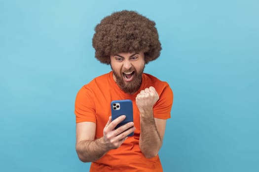 Portrait of extremely happy man with Afro hairstyle wearing orange T-shirt holding smart phone, looking at display, winning lottery, clenched fist. Indoor studio shot isolated on blue background.
